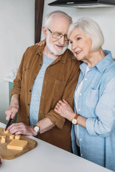 Sonriente esposa mayor abrazando marido corte queso en la tabla de cortar en la cocina - foto de stock