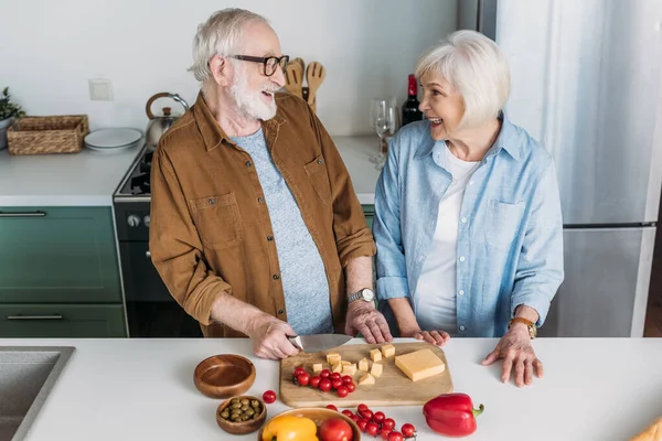 Happy elderly couple laughing while looking at each other near chopping board with cheese and vegetables in kitchen — Stock Photo