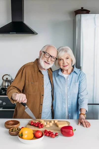 Feliz casal de idosos olhando para a câmera ao abraçar perto da mesa com comida na cozinha — Fotografia de Stock