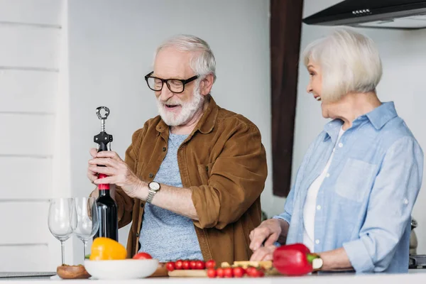 Souriant femme âgée regardant mari ouverture bouteille de vin avec tire-bouchon dans la cuisine sur le premier plan flou — Photo de stock