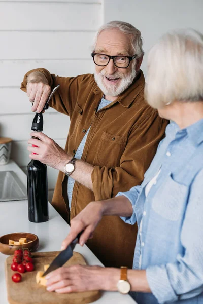 Heureux mari âgé regardant femme tout en ouvrant bouteille de vin avec tire-bouchon dans la cuisine sur le premier plan flou — Photo de stock