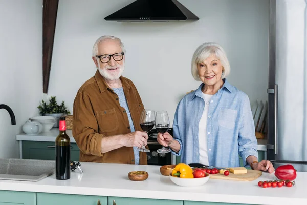 Heureux couple âgé regardant la caméra tout en grillant avec des verres à vin près de la table avec de la nourriture dans la cuisine — Photo de stock