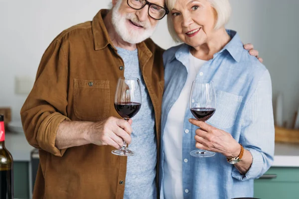 Sonriente pareja mayor abrazándose mientras sostiene copas de vino con cocina borrosa en el fondo - foto de stock