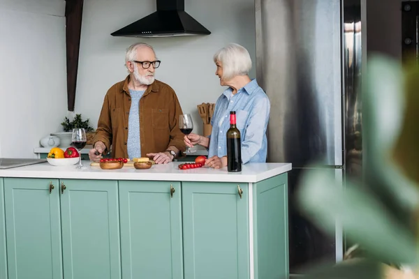 Hombre mayor mirando a la mujer con copa de vino mientras cocina la cena en la cocina en primer plano borrosa - foto de stock