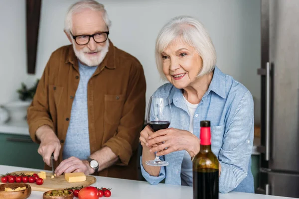 Sonriente marido mayor mirando a la esposa con copa de vino mientras se corta el queso en la tabla de cortar en la cocina sobre fondo borroso - foto de stock