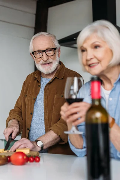Sonriente marido anciano mirando a la esposa con copa de vino mientras cocina la cena en la cocina en primer plano borrosa - foto de stock