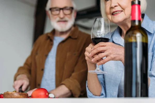 Donna anziana in possesso di bicchiere di vino vicino bottiglia con uomo sfocato su sfondo all'interno — Foto stock