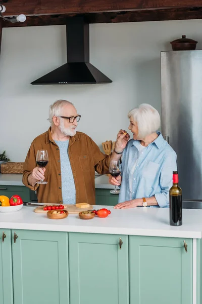 Sonriente anciano marido con copa de vino alimentación esposa con pedazo de queso en la cocina - foto de stock