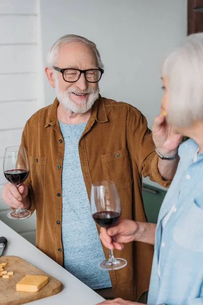 Happy elderly husband with wine glass feeding wife with piece of cheese in kitchen on blurred foreground — Stock Photo