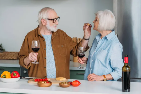 Smiling senior man with wine glass feeding wife with piece of cheese in kitchen on blurred background — Stock Photo