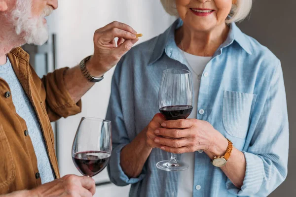 Cropped view of elderly husband with glass of wine feeding wife with piece of cheese in kitchen — Stock Photo