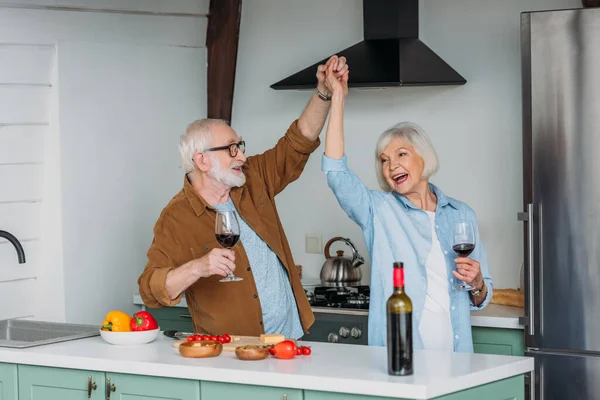 Feliz pareja de ancianos con copas de vino que se divierten cerca de la mesa con comida en la cocina - foto de stock