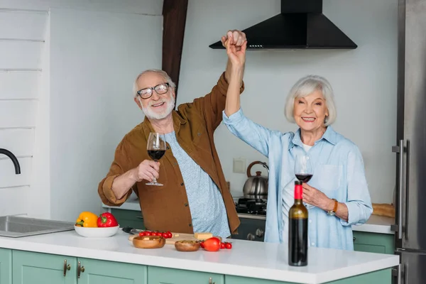 Glückliches Seniorenpaar mit Weingläsern, das in die Kamera schaut, während es sich am Tisch mit Essen in der Küche amüsiert — Stockfoto