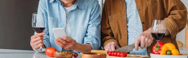 Ausgeschnittene Ansicht eines älteren Ehemanns, der in der Küche mit Weinglas und Smartphone in der Nähe seiner Frau Abendessen kocht, Banner — Stockfoto