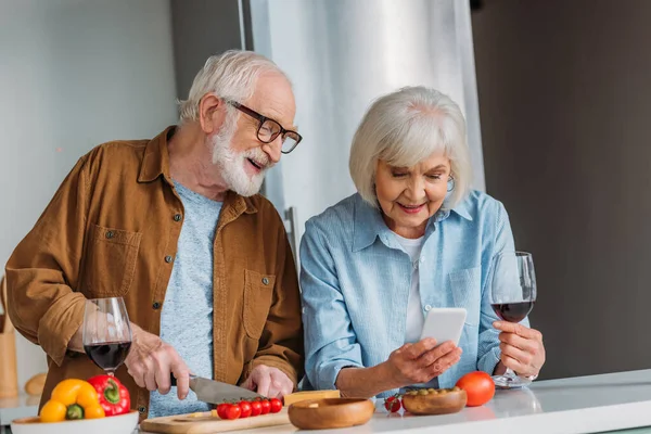 Glücklicher Senior-Ehemann schaut beim Kochen in der Küche auf Smartphone neben Ehefrau — Stockfoto