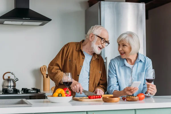 Feliz pareja de ancianos mirándose cerca de la mesa con comida y copas de vino en la cocina - foto de stock