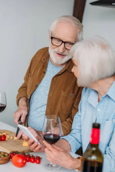 Smiling senior husband looking at wife while cutting cheese on chopping board near vegetables on blurred foreground — Stock Photo