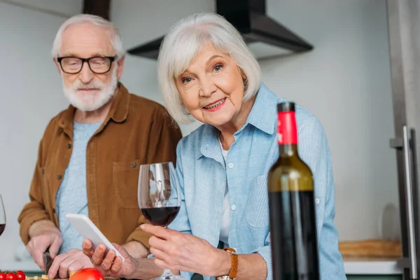 Lächelnde Seniorin mit Smartphone und Weinglas, die mit verschwommener Flasche im Vordergrund in die Kamera schaut — Stockfoto