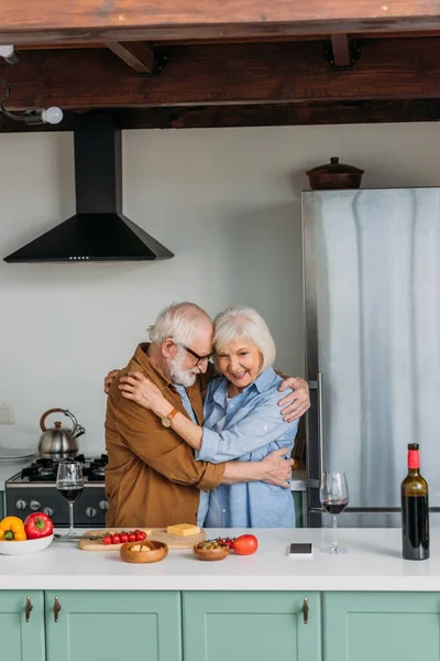 Felice coppia anziana che si abbraccia vicino al tavolo con bicchieri di cibo e vino in cucina — Foto stock