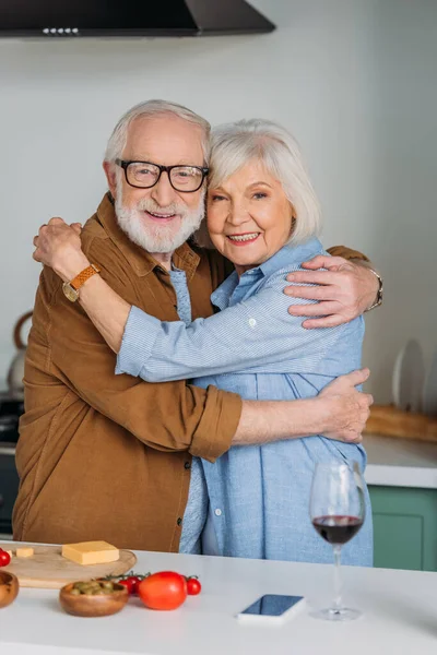 Felice coppia anziana guardando la fotocamera mentre abbraccia vicino al tavolo con cibo in cucina su sfondo sfocato — Foto stock