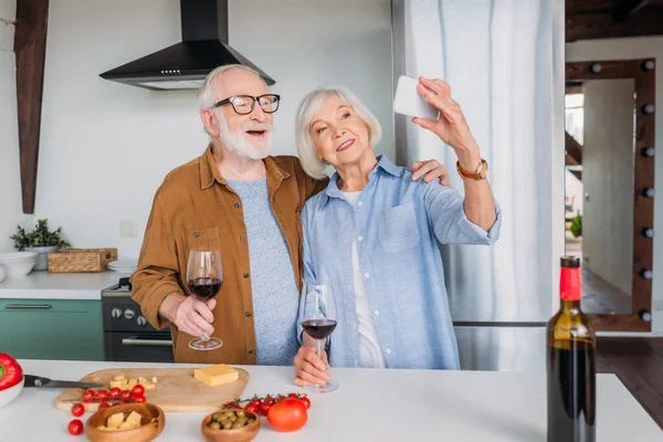 Feliz casal de idosos com copos de vinho tomando selfie perto da mesa com comida na cozinha — Fotografia de Stock
