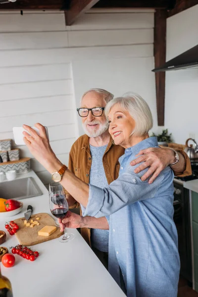 Sorrindo casal de idosos com copos de vinho abraçando ao tomar selfie perto da mesa com comida na cozinha — Fotografia de Stock