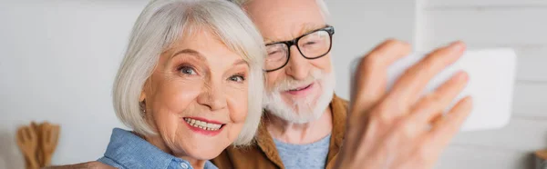 Joyeuse femme âgée regardant la caméra tout en prenant selfie avec l'homme sur le premier plan flou à l'intérieur, bannière — Photo de stock