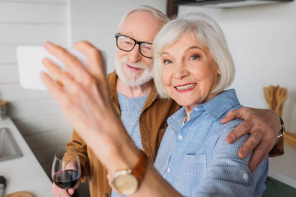 Heureuse femme âgée regardant la caméra tout en prenant selfie avec mari tenant verre de vin à l'intérieur sur le premier plan flou — Photo de stock