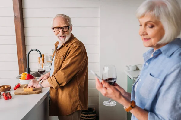 Homme âgé souriant regardant la femme tout en coupant du fromage sur la planche à découper dans la cuisine sur le premier plan flou — Photo de stock