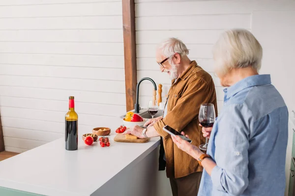 Літня дружина зі смартфоном і келихом вина біля усміхненого чоловіка готує вечерю на кухні на розмитому фоні — стокове фото