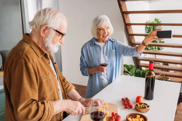Heureuse femme âgée avec verre à vin prendre selfie près de mari cuisine dîner dans la cuisine — Photo de stock