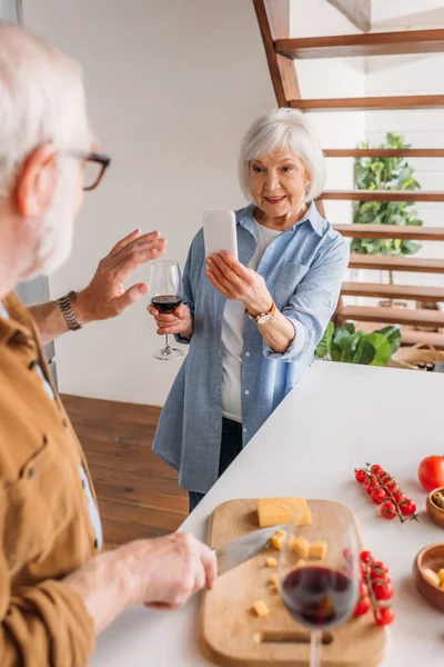 Femme âgée souriante avec smartphone prenant la photo du mari en train de cuisiner le dîner au premier plan flou dans la cuisine — Photo de stock