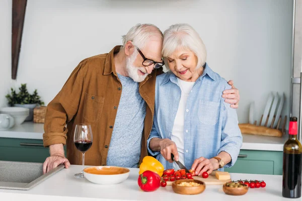 Mari aîné souriant étreignant femme cuisine le dîner sur la table avec du vin, du fromage et des légumes sur fond flou — Photo de stock