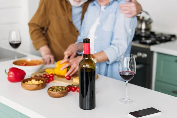 Cropped view of elderly couple near table with bottle of wine and vegetarian food on blurred background — Stock Photo