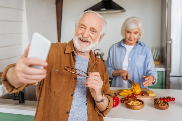 Щасливий старший чоловік зі смартфоном, який приймає селфі з дружиною готує вечерю на кухні на розмитому фоні — стокове фото