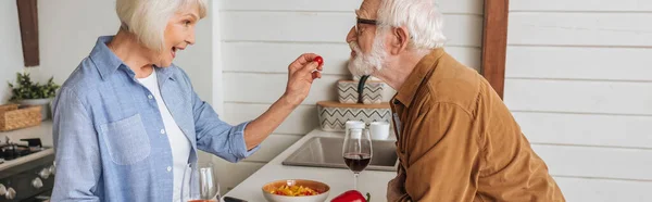 Seitenansicht der glücklichen älteren Frau, die ihren Mann mit Kirschtomaten in der Küche füttert, Banner — Stockfoto