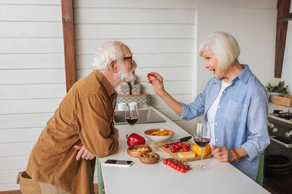 Side view of happy senior wife feeding husband with cherry tomato near table with food, wine glasses and smartphone in kitchen — Stock Photo