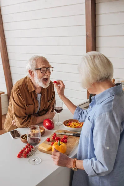 Vue arrière de la femme aînée nourrir mari heureux avec tomate cerise près de la table avec de la nourriture dans la cuisine — Photo de stock