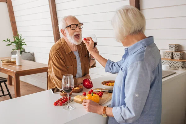 Visão traseira da esposa idosa alimentando marido feliz com tomate cereja perto da mesa com comida na cozinha — Fotografia de Stock