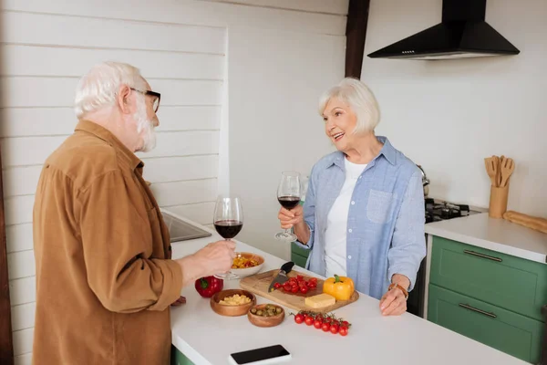 Sonriente hombre mayor con copa de vino mirando al marido cerca de la mesa con comida en la cocina - foto de stock