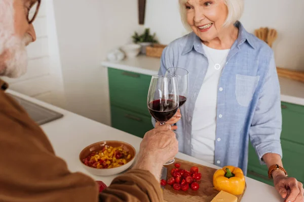 Happy elderly couple toasting with wine glasses near table with food on blurred foreground — Stock Photo