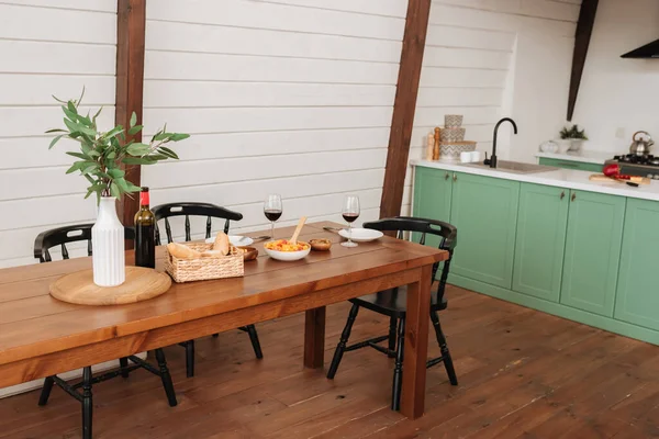 Mesa servida con cena vegetariana, botella de vino y copas cerca de planta decorativa en jarrón en cocina - foto de stock