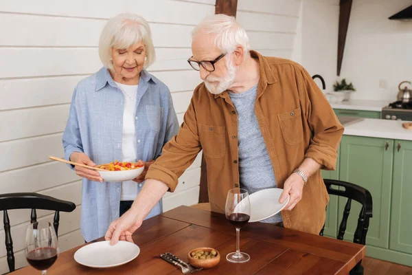 Lächelndes Seniorenpaar serviert Tisch mit Tellern und Salat in Küche auf verschwommenem Hintergrund — Stockfoto