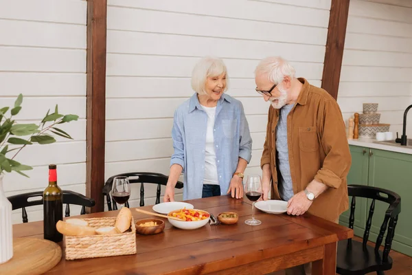 Feliz pareja de ancianos sirviendo mesa con platos en la cocina - foto de stock