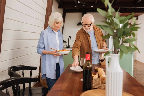 Sonriente pareja de ancianos sirviendo mesa con platos y ensalada en la cocina con planta decorativa borrosa en primer plano - foto de stock