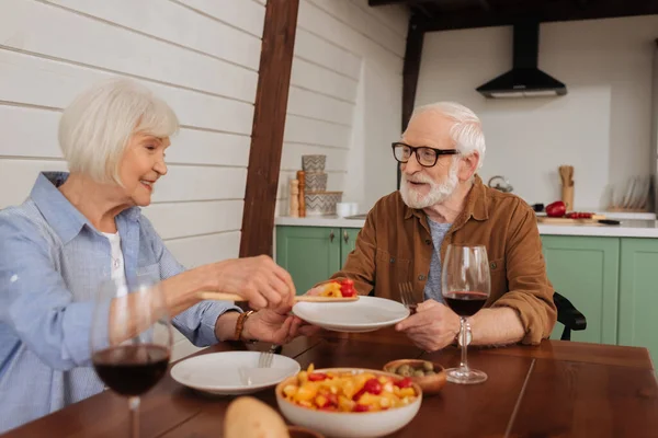 Sorridente moglie anziana con spatola che serve insalata per marito a tavola con cena in primo piano sfocata — Foto stock