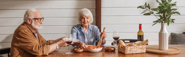 Sonriente esposa mayor con espátula que sirve ensalada para el marido en la mesa con cena vegetariana en la cocina, pancarta - foto de stock