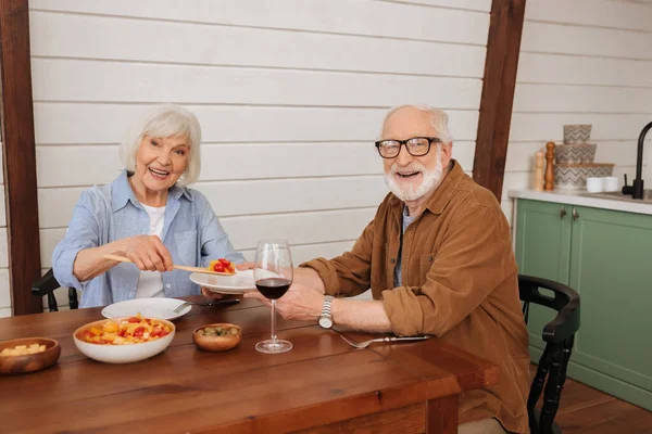 Sonriente esposa mayor mirando a la cámara mientras sirve ensalada para el marido en la mesa con cena vegetariana en la cocina - foto de stock