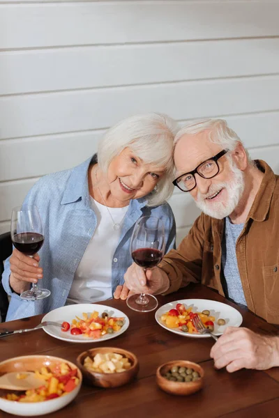 Glückliches Seniorenpaar mit Weingläsern, die in die Kamera schauen, während sie am Tisch sitzen und vegetarisches Abendessen im verschwommenen Vordergrund genießen — Stockfoto