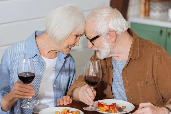 Feliz pareja de ancianos con copas de vino mirándose mientras están sentados en la mesa con la cena vegetariana en un fondo borroso — Stock Photo